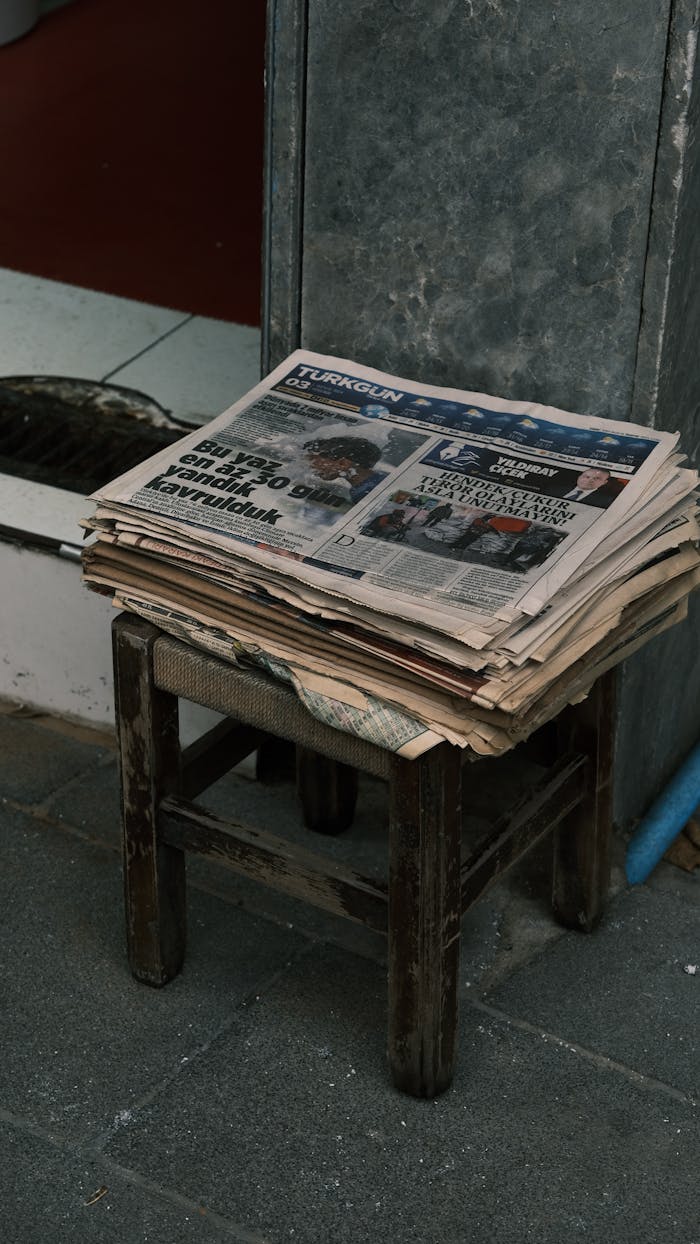 Old newspapers stacked on a wooden stool outdoors on urban street corner.